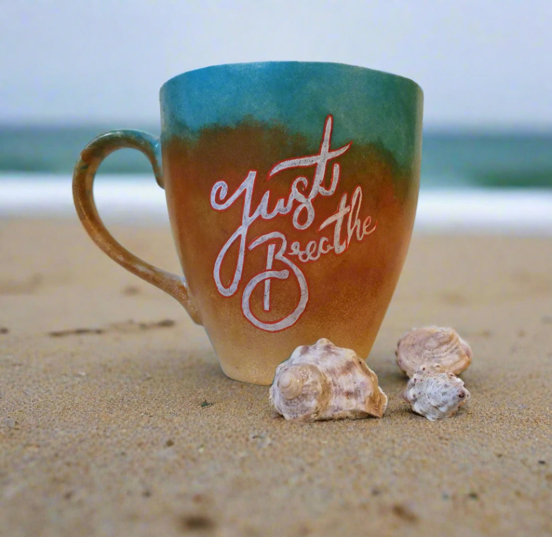 A gradient colored hand painted mug (from sand color at the botom to the sea color on top) with the words "Just breathe""