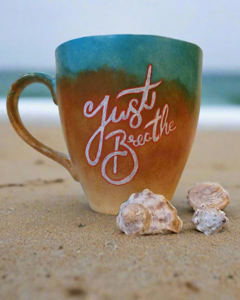 A gradient colored hand painted mug (from sand color at the botom to the sea color on top) with the words "Just breathe""