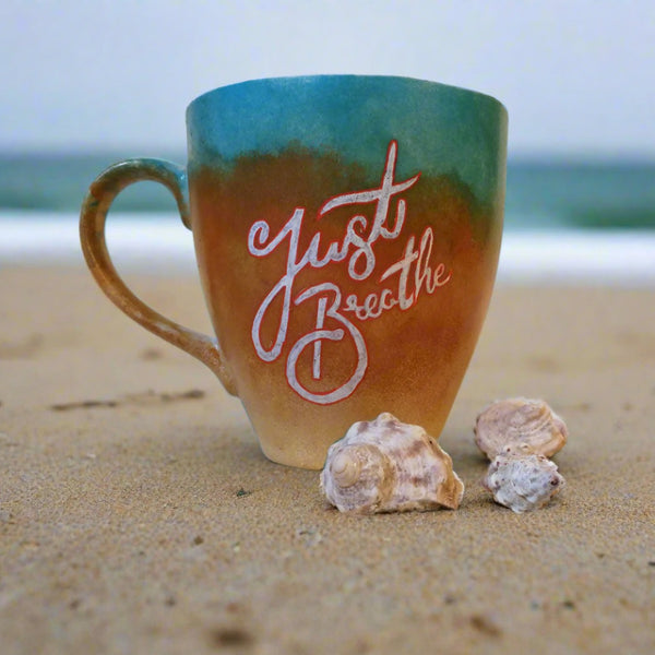 A gradient colored hand painted mug (from sand color at the botom to the sea color on top) with the words "Just breathe""