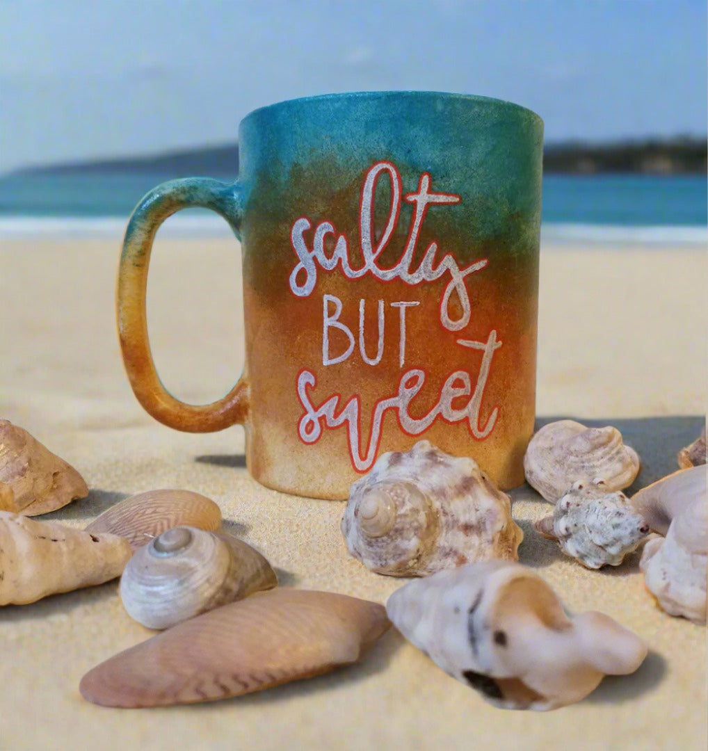 A gradient colored hand painted mug (from sand color at the bottom to the sea color on top) with the words "Salty but Sweet"