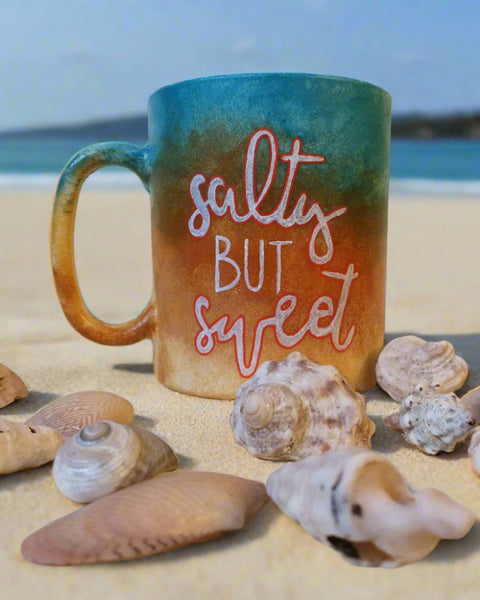 A gradient colored hand painted mug (from sand color at the bottom to the sea color on top) with the words "Salty but Sweet"