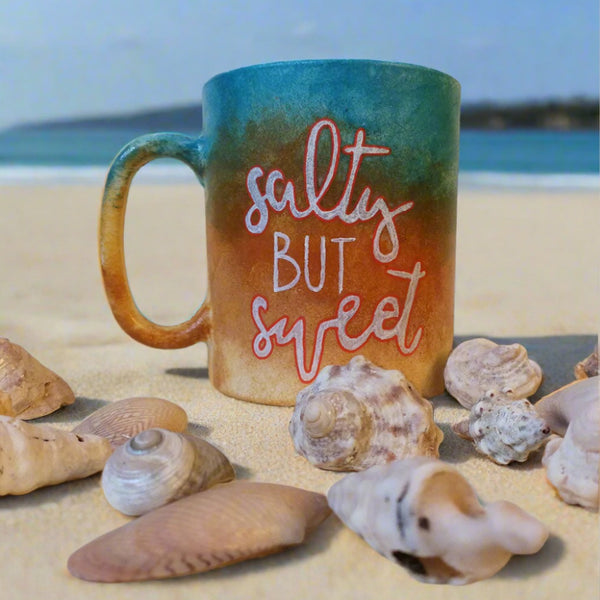 A gradient colored hand painted mug (from sand color at the bottom to the sea color on top) with the words "Salty but Sweet"