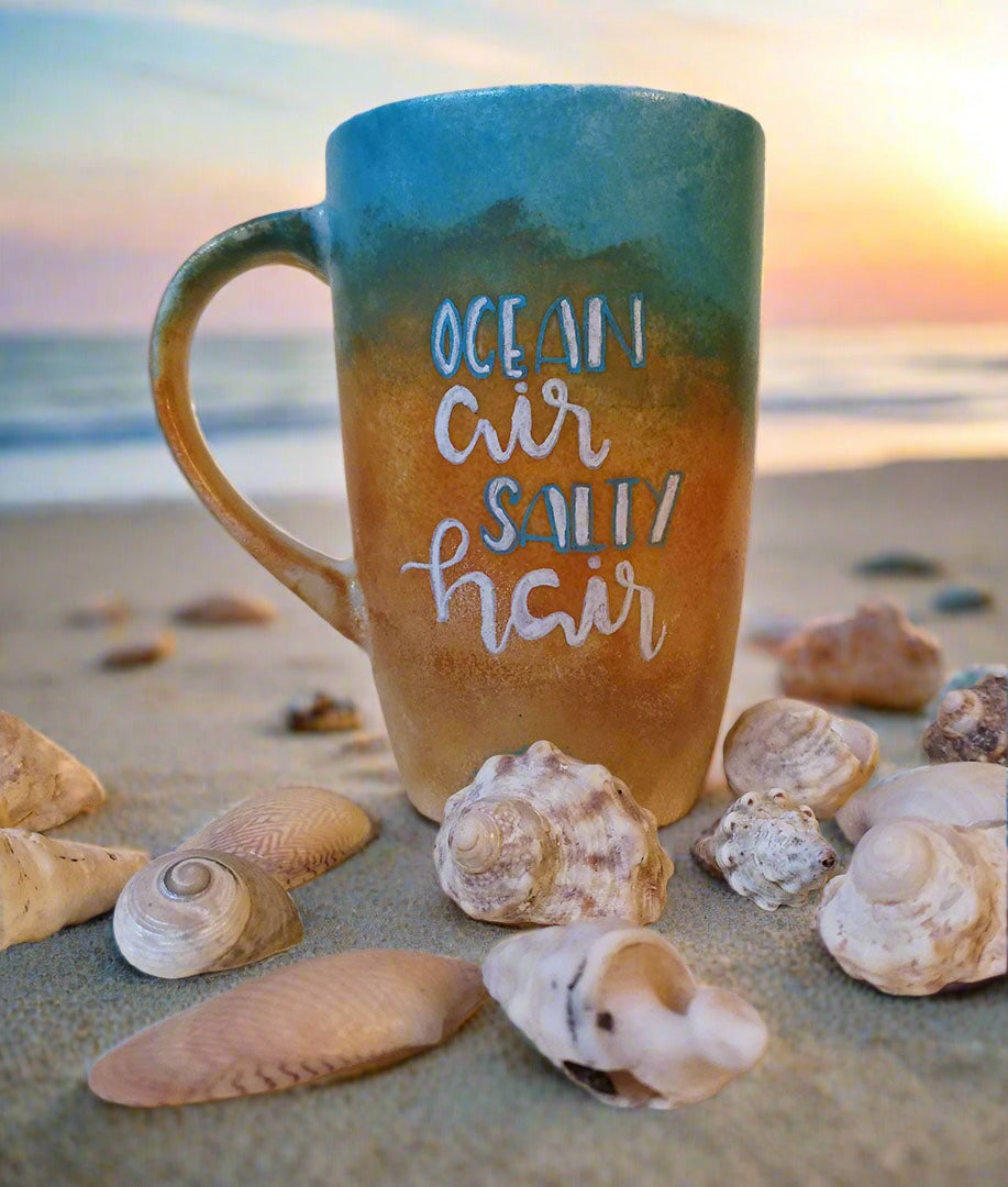 A gradient colored hand apinted mug (from sand color at the botom to the sea color on top) with the words "ocean air, salty hair"