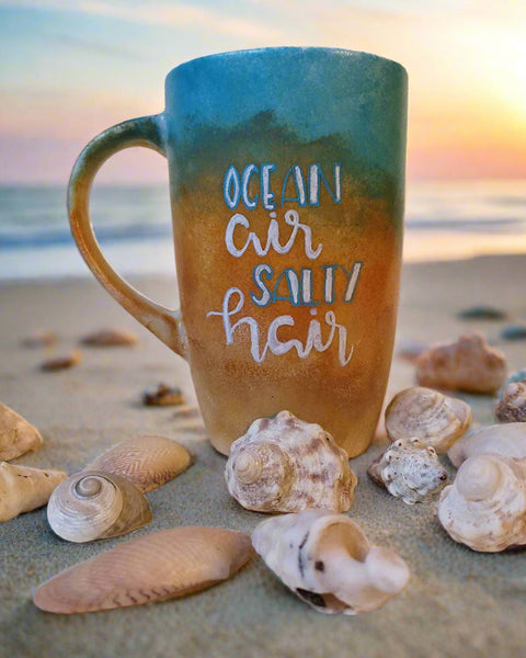 A gradient colored hand apinted mug (from sand color at the botom to the sea color on top) with the words "ocean air, salty hair"