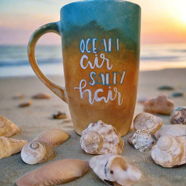 A gradient colored hand apinted mug (from sand color at the botom to the sea color on top) with the words "ocean air, salty hair"