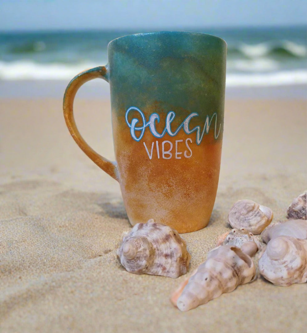 A gradient colored hand apinted mug (from sand color at the botom to the sea color on top) with the words "Ocean Vibes"