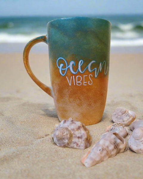 A gradient colored hand apinted mug (from sand color at the botom to the sea color on top) with the words "Ocean Vibes"