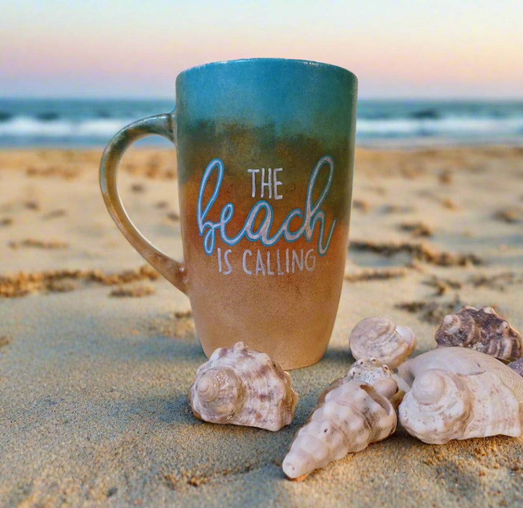 A gradient colored hand painted mug (from sand color at the botom to the sea color on top) with the words "the beach is calling"