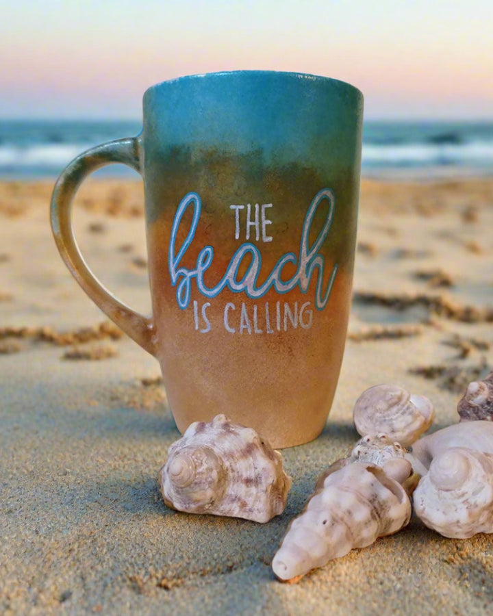 A gradient colored hand painted mug (from sand color at the botom to the sea color on top) with the words "the beach is calling"