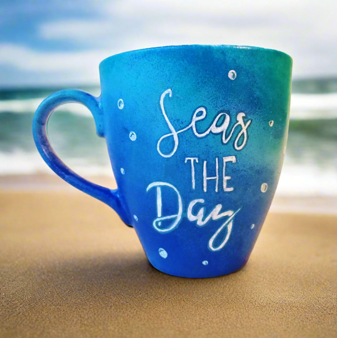 A Bluish hand painted mug with the words "Seas the Day" written in blue and colored with white, and bubbles scattered all over