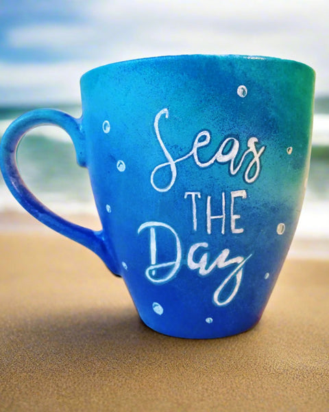 A Bluish hand painted mug with the words "Seas the Day" written in blue and colored with white, and bubbles scattered all over