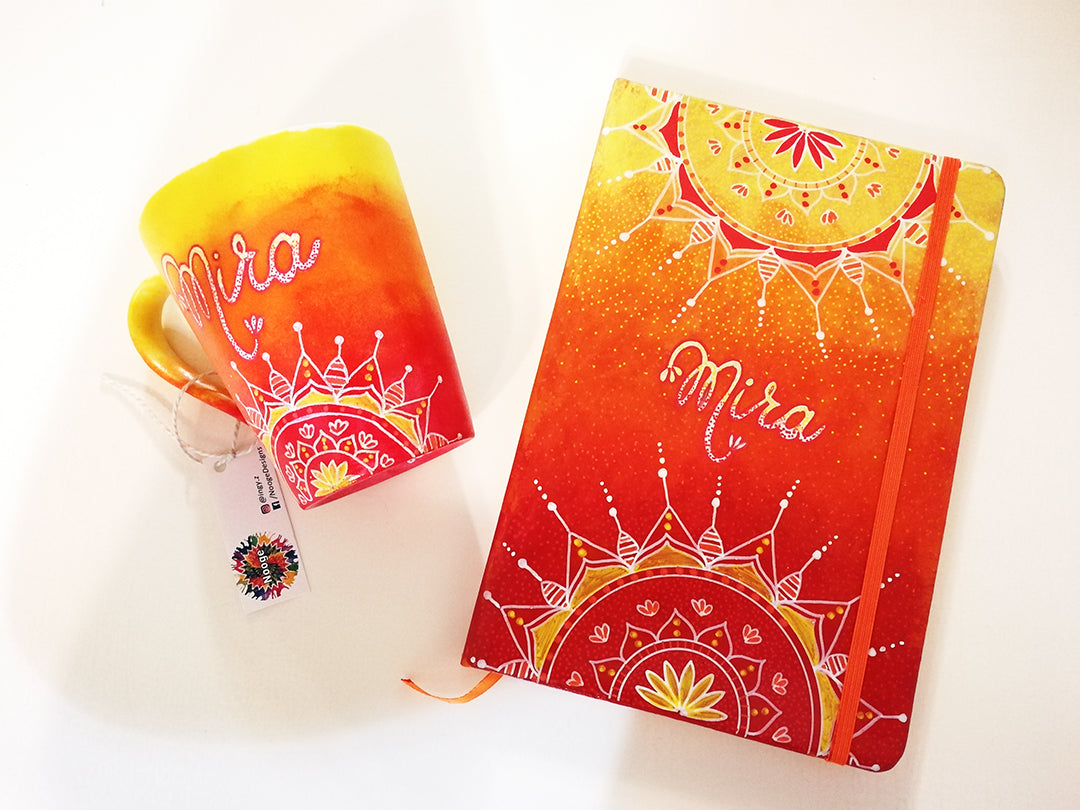 A Gift Set  hand apinted in gradient colors (yellow-orange-red) with the mug hacving  half mandala on the bottom and the name mira in the center and the notebook having half mandalas on both the top and bottom and the name mira in the center