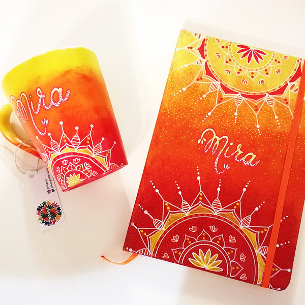 A Gift Set  hand apinted in gradient colors (yellow-orange-red) with the mug hacving  half mandala on the bottom and the name mira in the center and the notebook having half mandalas on both the top and bottom and the name mira in the center