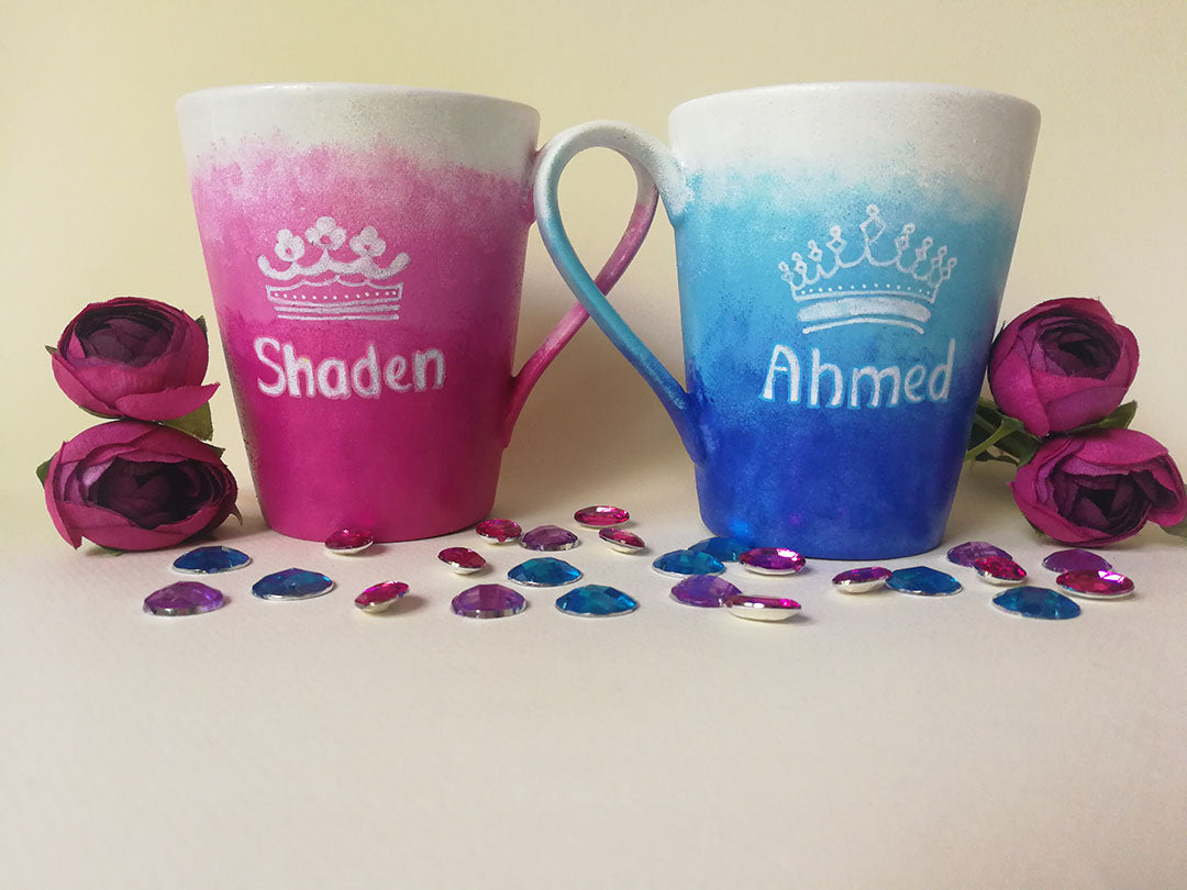 2 hand-painted mugs. One with gradients of pink and has the name "Shaden" and a crown drawn above the name and the other is colored in gradients of blue and has the name "Ahmed" and a crown drawn above the name