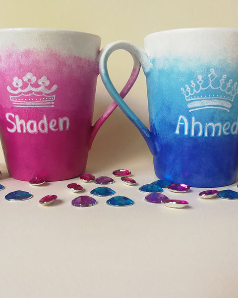 2 hand-painted mugs. One with gradients of pink and has the name "Shaden" and a crown drawn above the name and the other is colored in gradients of blue and has the name "Ahmed" and a crown drawn above the name