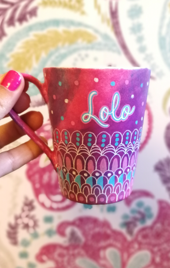 A Pink hand painted mug with patterns on the bottom half horizontally in white and colored in pink and blue with the name "LoLo" on the top