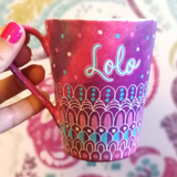 A Pink hand painted mug with patterns on the bottom half horizontally in white and colored in pink and blue with the name 