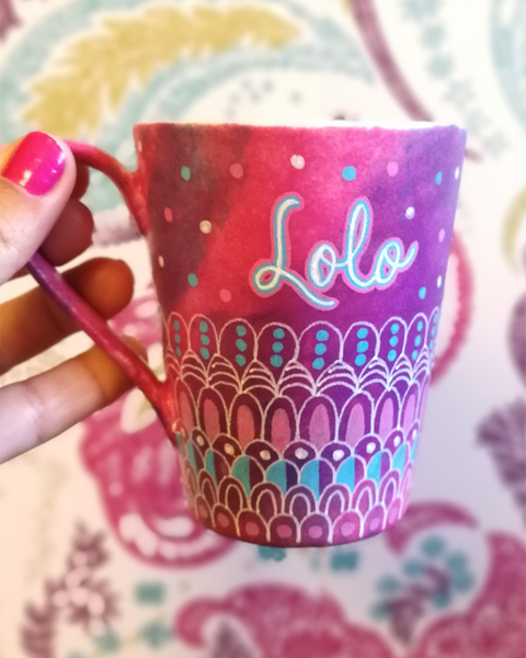 A Pink hand painted mug with patterns on the bottom half horizontally in white and colored in pink and blue with the name "LoLo" on the top