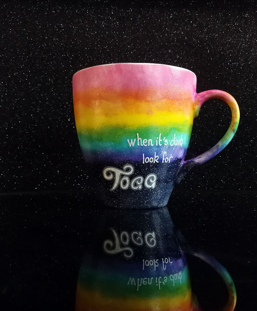 A Mug with the top half painted in rainbow and the bottom in dark galaxy, with the name on the dark galaxy "Toaa"