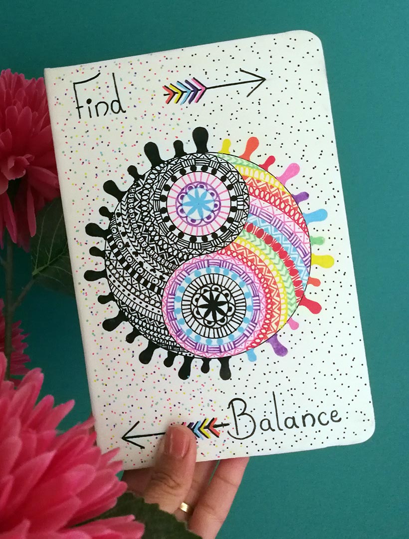 A Pearl white hand painted notebook with a yin and yang stuck together in a circle drawn in the center with one side totally in black patterns and the other in patterns of different colors, with the word "Find" written on the top and an arrow pointing to the right. and the word 'Balance" written at the bottom and an arrow pointing to the left