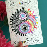 A Pearl white hand painted notebook with a yin and yang stuck together in a circle drawn in the center with one side totally in black patterns and the other in patterns of different colors, with the word 