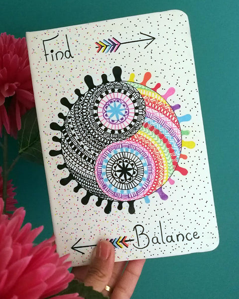 A Pearl white hand painted notebook with a yin and yang stuck together in a circle drawn in the center with one side totally in black patterns and the other in patterns of different colors, with the word "Find" written on the top and an arrow pointing to the right. and the word 'Balance" written at the bottom and an arrow pointing to the left