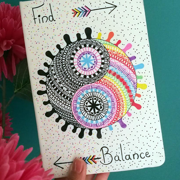 A Pearl white hand painted notebook with a yin and yang stuck together in a circle drawn in the center with one side totally in black patterns and the other in patterns of different colors, with the word "Find" written on the top and an arrow pointing to the right. and the word 'Balance" written at the bottom and an arrow pointing to the left