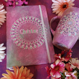 A gift set of mug and notebook in pastel pink , both with mandalas. and the notebook has the name christine inside the mandala