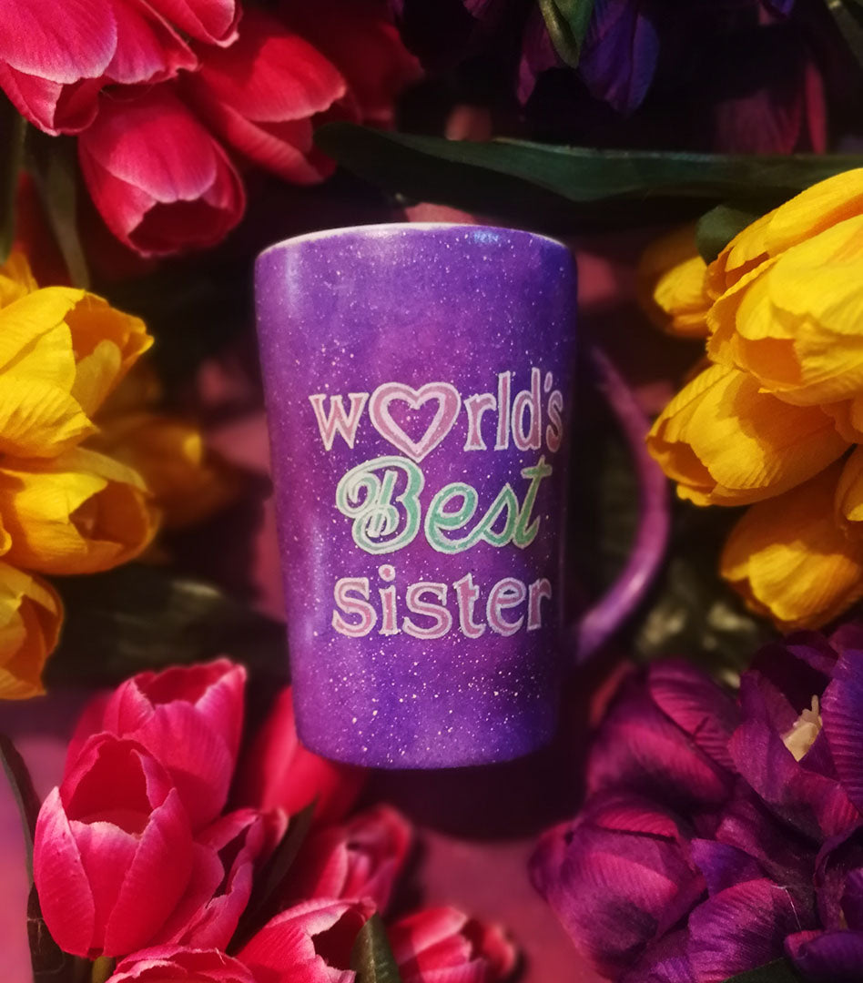 A Purple galaxy hand painted mug with the sentence "Worlds best sister" written in white and colored in with pink and green