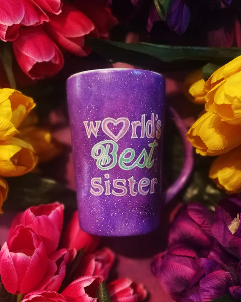 A Purple galaxy hand painted mug with the sentence "Worlds best sister" written in white and colored in with pink and green