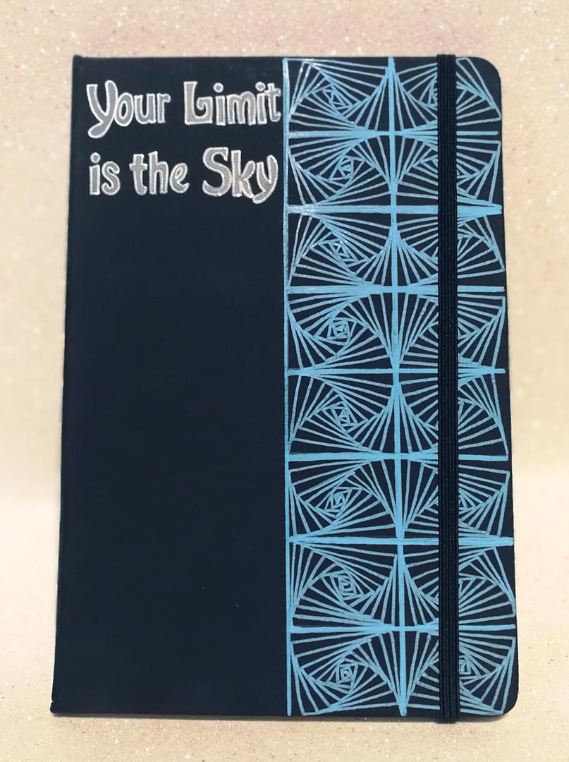 A Dark Blue Notebook with patterns drawn Vertically on half of the notebook top down and the the half has the words "your limit is the sky"