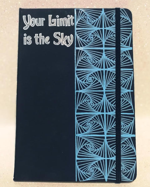 A Dark Blue Notebook with patterns drawn Vertically on half of the notebook top down and the the half has the words "your limit is the sky"