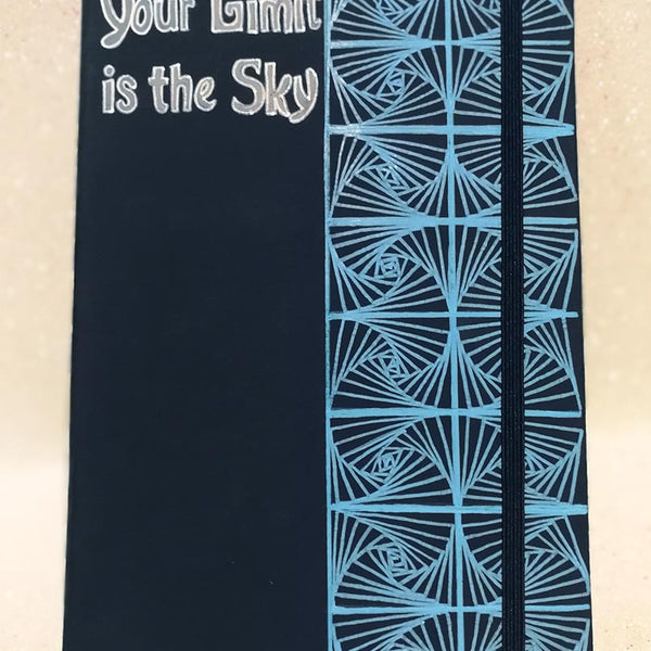 A Dark Blue Notebook with patterns drawn Vertically on half of the notebook top down and the the half has the words "your limit is the sky"