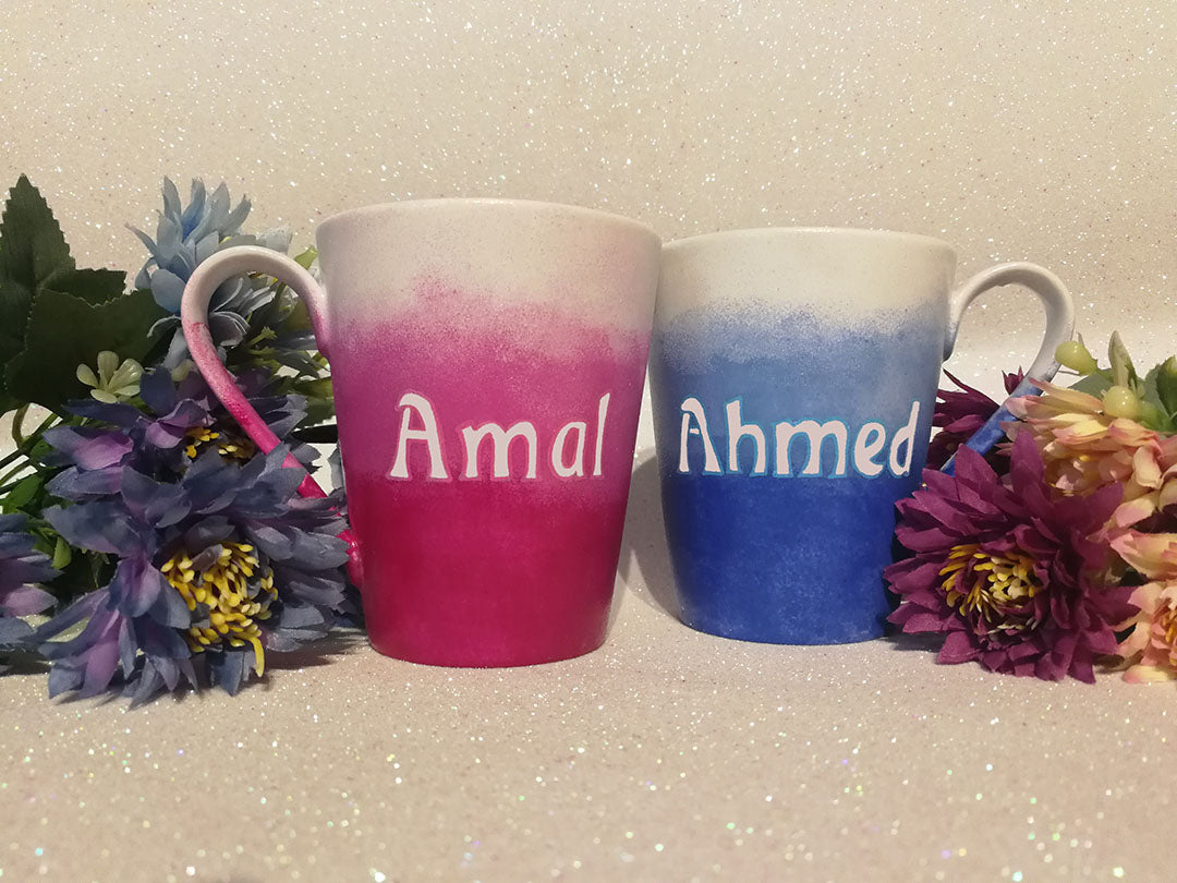 2 hand-painted mugs. one colored in gradient blues to white and has the name "ahmed". the other is colored in gradient pinks to white and has the name "Amal" on it