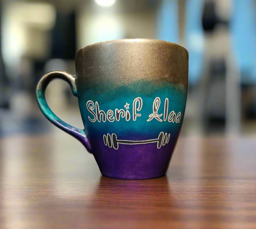 A gradient colored mug (Silver, turquoise and purple) with the name "Sherif Alaa"and a small weight bar drawn below it