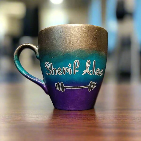 A gradient colored mug (Silver, turquoise and purple) with the name "Sherif Alaa"and a small weight bar drawn below it