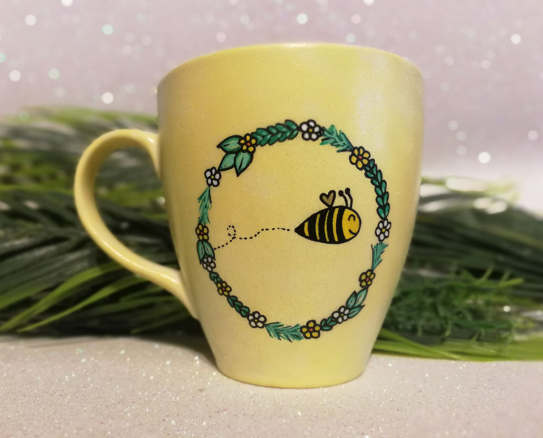 A pastel yellow mug with a flower wreth and a bee inside it