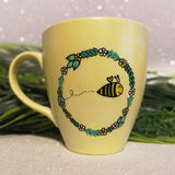 A pastel yellow mug with a flower wreth and a bee inside it