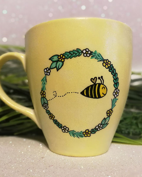 A pastel yellow mug with a flower wreth and a bee inside it