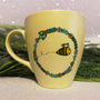 A pastel yellow mug with a flower wreth and a bee inside it