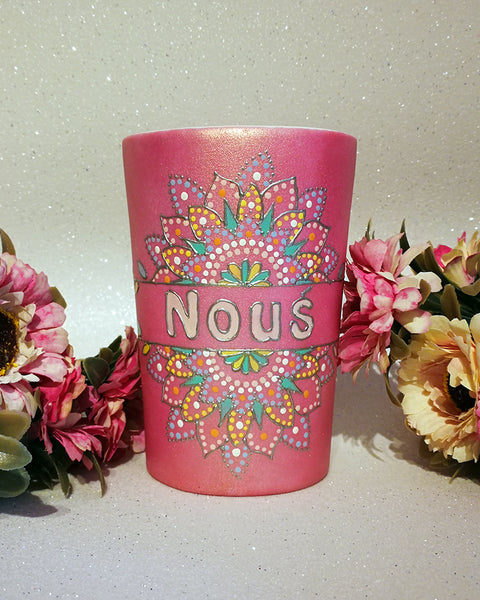 A Shimmery pink hand painted mug with a mandala split into 2 with a space in between that has the name "Nous" and the mandala is outlined with silver relief and dot art of white, baby pink, mint green, yellow and baby blue