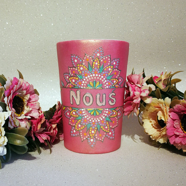 A Shimmery pink hand painted mug with a mandala split into 2 with a space in between that has the name "Nous" and the mandala is outlined with silver relief and dot art of white, baby pink, mint green, yellow and baby blue