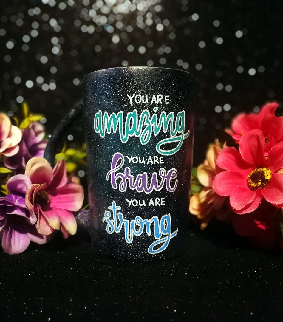 A Black galaxy hand painted mug with the words "you are amazing, you are brave, you are strong" with the words Amazing brave and strong in larger cursive text