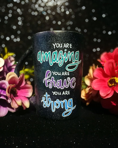 A Black galaxy hand painted mug with the words "you are amazing, you are brave, you are strong" with the words Amazing brave and strong in larger cursive text