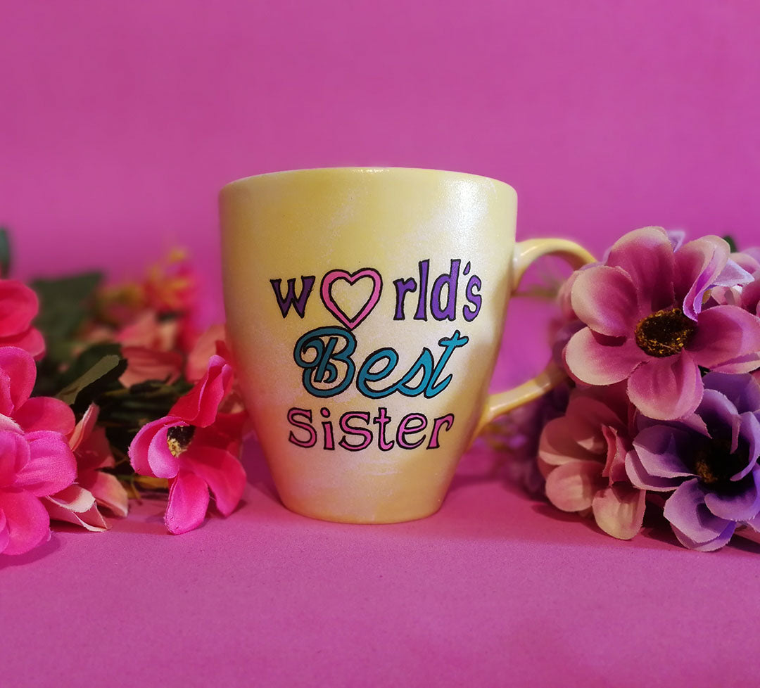 A Pastel yellow hand painted mug with the sentence "Worlds best Sister" written in black and colored in purple , pink and turquoise