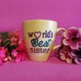 A Pastel yellow hand painted mug with the sentence "Worlds best Sister" written in black and colored in purple , pink and turquoise