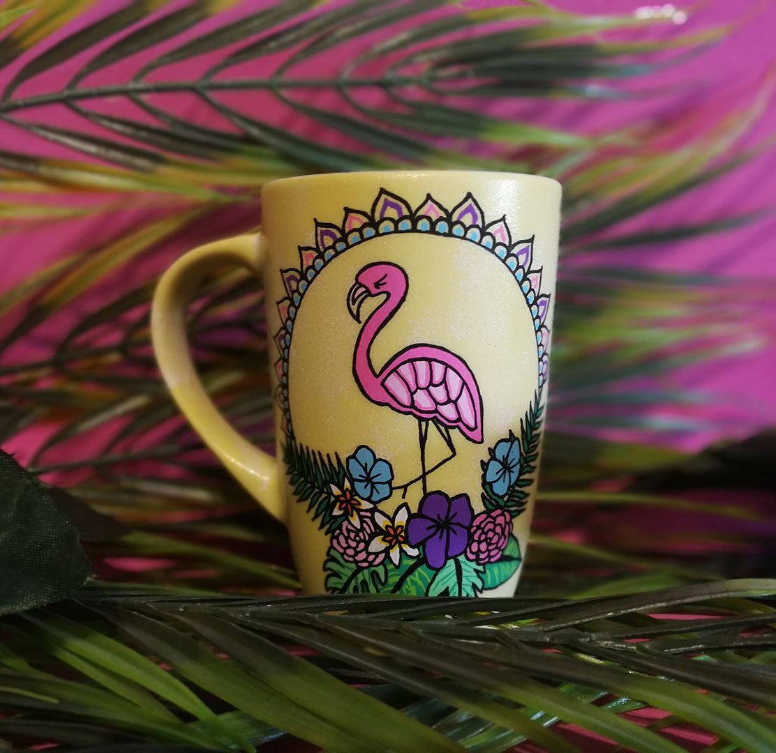 A Pastel yellow hand painted mug with a flamingo standing in the center over colorful flowers and around it half a mandala wreth