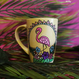 A Pastel yellow hand painted mug with a flamingo standing in the center over colorful flowers and around it half a mandala wreth