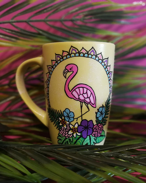 A Pastel yellow hand painted mug with a flamingo standing in the center over colorful flowers and around it half a mandala wreth