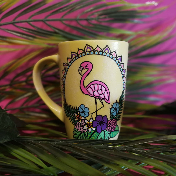 A Pastel yellow hand painted mug with a flamingo standing in the center over colorful flowers and around it half a mandala wreth